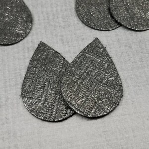 May include: Close-up of several silver teardrop-shaped earrings with a textured, metallic finish. The earrings are arranged on a light gray surface. The image showcases the jewelry's design and material.