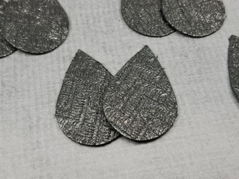 May include: Close-up of several silver teardrop-shaped earrings with a textured, metallic finish. The earrings are arranged on a light gray surface. The image showcases the jewelry's design and material.