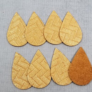 May include: Eight teardrop-shaped leather earring blanks. Seven are a textured gold color with a woven pattern, and one is a solid brown suede. The earrings are arranged on a gray surface.