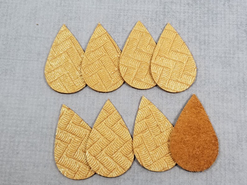 May include: Eight teardrop-shaped leather earring blanks. Seven are a textured gold color with a woven pattern, and one is a solid brown suede. The earrings are arranged on a gray surface.