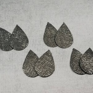 May include: Six pairs of silver teardrop-shaped earrings with a textured surface are arranged on a light gray background. The earrings are made of a metallic material and have a subtle, woven pattern.