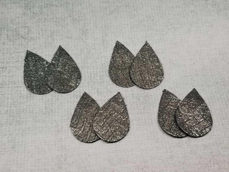 May include: Six pairs of silver teardrop-shaped earrings with a textured surface are arranged on a light gray background. The earrings are made of a metallic material and have a subtle, woven pattern.