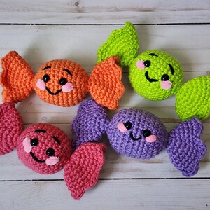May include: Crocheted candy toys in orange, pink, green, and purple. Each candy has a smiling face with pink cheeks and black eyes. The candies are arranged on a light wood surface.