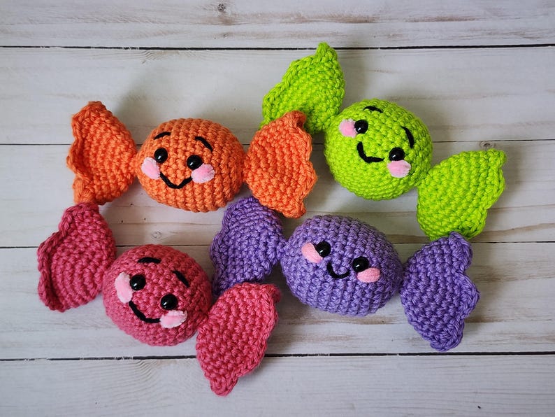 May include: Crocheted candy toys in orange, pink, green, and purple. Each candy has a smiling face with pink cheeks and black eyes. The candies are arranged on a light wood surface.