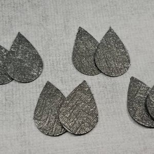 May include: Six pairs of silver teardrop-shaped earrings with a textured, woven pattern. The earrings are arranged on a light gray surface. The earrings are likely for pierced ears and are a neutral color.
