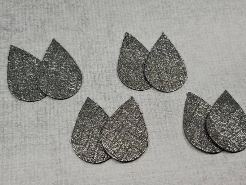 May include: Six pairs of silver teardrop-shaped earrings with a textured, woven pattern. The earrings are arranged on a light gray surface. The earrings are likely for pierced ears and are a neutral color.