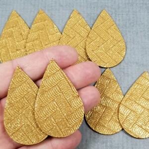 May include: Gold teardrop-shaped faux leather earring blanks with a textured, woven pattern. Several blanks are visible, some held in a hand. The earrings are a metallic gold color.