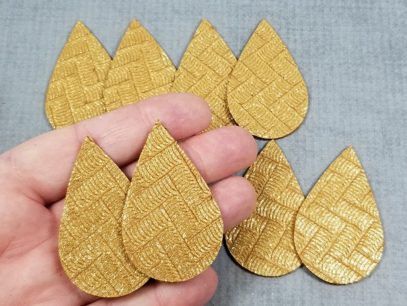 May include: Gold teardrop-shaped faux leather earring blanks with a textured, woven pattern. Several blanks are visible, some held in a hand. The earrings are a metallic gold color.