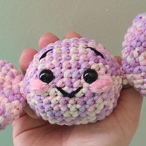 May include: A handmade crochet candy with a smiling face and two wings. The candy is made of pink, purple, and white yarn. It has black eyes and pink cheeks. The candy is held in a hand.