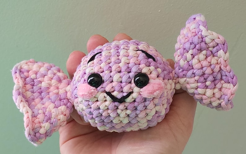 May include: A handmade crochet candy with a smiling face and two wings. The candy is made of pink, purple, and white yarn. It has black eyes and pink cheeks. The candy is held in a hand.