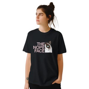 Könnte beinhalten: Schwarzes T-Shirt mit einer Grafik einer grummeligen Katze und dem rosa Text "THE NOPE FACE". Die Katze hat ein weißes Gesicht, braune Ohren und ein blaues Auge. Das T-Shirt ist ein klassischer Rundhalsausschnitt.