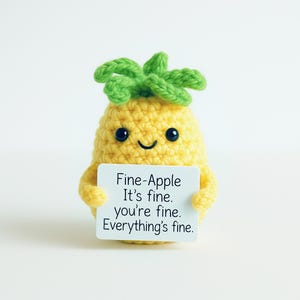May include: A yellow crocheted pineapple with green leaves and black eyes holds a white sign. The sign reads "Fine-Apple It's fine. you're fine. Everything's fine."