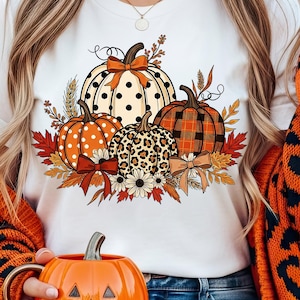 May include: White t-shirt featuring a fall-themed pumpkin design. The design includes pumpkins with polka dots, leopard print, and plaid patterns, accented with bows and fall foliage. The person is wearing an orange sweater with black leopard print.