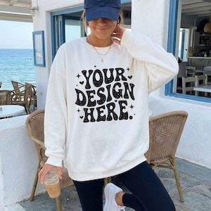 May include: A white crewneck sweatshirt with the text "YOUR DESIGN HERE" in a black, retro font. The sweatshirt is worn by a person wearing a blue baseball cap and black leggings. The person is holding a clear cup with a brown beverage.