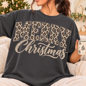 Könnte beinhalten: Ein anthrazitfarbenes Kurzarm-T-Shirt mit dem Schriftzug MERRY in Blockbuchstaben mit braun-schwarzem Leopardenmuster und dem Wort Christmas in beiger Schreibschrift auf der Brust.