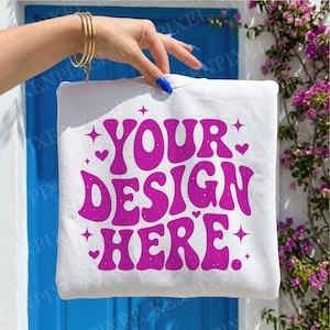 May include: White sweatshirt with the text "YOUR DESIGN HERE" in a retro, bubble font. The text is in a vibrant magenta color, with hearts and star accents. The sweatshirt is held up by a hand with gold bracelets and blue nail polish.