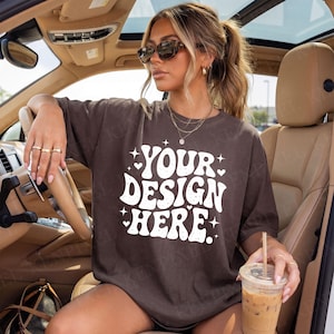 Pode incluir: Uma camiseta marrom com o texto "YOUR DESIGN HERE" em branco, com detalhes de estrelas e corações. A camiseta é usada por uma pessoa em um carro, segurando uma bebida. A camiseta é uma tela em branco para designs personalizados.