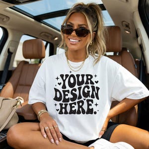 以下が含まれることがあります： 黒いレトロフォントで「YOUR DESIGN HERE」というテキストが入った白いTシャツ。 サングラスと金のジュエリーを身に着けた人がシャツを着ています。 その人は茶色の内装の車の中に座っています。