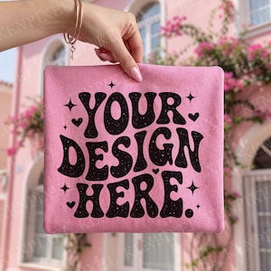 以下が含まれることがあります： ピンク色の正方形の布に、黒のレトロフォントで「YOUR DESIGN HERE」と書かれています。星とハートの装飾が施されています。 金色のブレスレットをつけた手で布が持たれています。