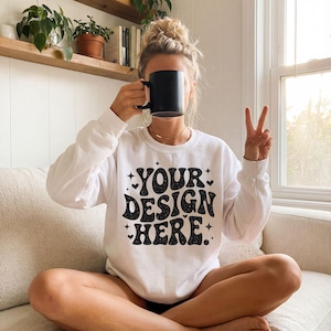 May include: White long-sleeve sweatshirt with the text "YOUR DESIGN HERE" in a retro font. The sweatshirt is being held by a person holding a black mug. The person is sitting on a beige couch.