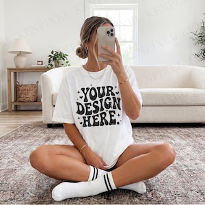 Pode incluir: Camiseta branca com o texto "YOUR DESIGN HERE" em uma fonte preta retr&ocirc;. A camiseta &eacute; usada com meias brancas com listras pretas. A pessoa est&aacute; sentada em um tapete estampado em um quarto bem iluminado.
