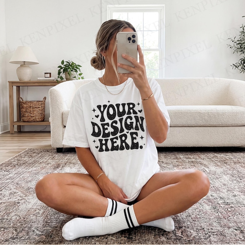 Pode incluir: Camiseta branca com o texto "YOUR DESIGN HERE" em uma fonte preta retr&ocirc;. A camiseta &eacute; usada com meias brancas com listras pretas. A pessoa est&aacute; sentada em um tapete estampado em um quarto bem iluminado.