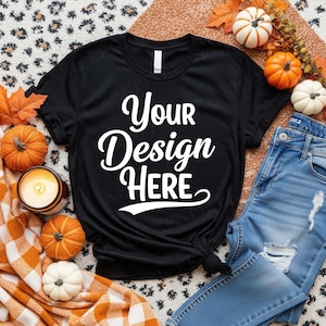 May include: Black t-shirt with the text "Your Design HERE" in white script. The shirt is styled with blue jeans, pumpkins, fall leaves, and a lit candle, suggesting a seasonal or autumn theme.