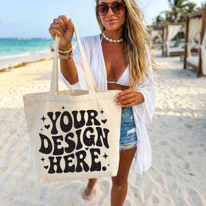 May include: A cream-coloured tote bag featuring the text "YOUR DESIGN HERE" in black, decorative lettering. The bag has long handles. The background is a beach scene.