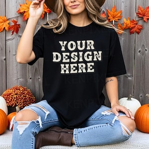 Pode incluir: Camiseta preta com o texto "YOUR DESIGN HERE" em uma fonte de estilo vintage. A modelo está usando jeans azuis rasgados e um chapéu marrom. O fundo inclui abóboras, folhagens de outono e uma parede de madeira.