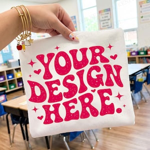 May include: White t-shirt with the text "YOUR DESIGN HERE" in a retro, bubble font. The text is in a bright pink color, with hearts and starbursts. The shirt is being held up in a classroom setting.