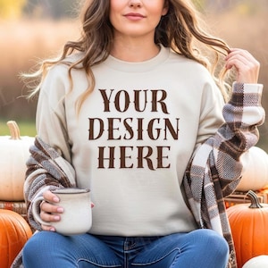 May include: Beige sweatshirt with the text "YOUR DESIGN HERE" in brown lettering. The person is wearing a brown and white plaid shawl and holding a beige mug. Pumpkins are in the background.