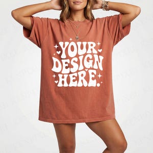 Puede incluir: Una camiseta de color óxido con el texto blanco "YOUR DESIGN HERE" en un estilo retro. La camiseta de manga corta tiene un corte holgado, adecuada para ropa casual. El diseño incluye estrellas y corazones.