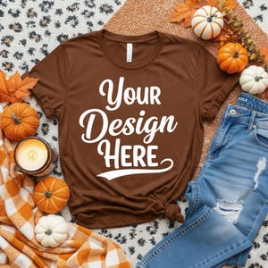 May include: A brown t-shirt with the text "Your Design Here" in white script. The shirt is styled with pumpkins, fall leaves, a candle, and blue jeans. The scene evokes a cozy autumn theme.