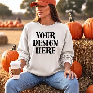 May include: A white sweatshirt with the text "YOUR DESIGN HERE" in black, paired with blue jeans and an orange baseball cap. The scene includes pumpkins and a coffee cup, suggesting a fall theme.