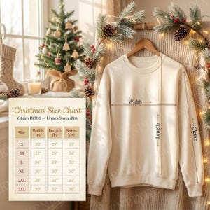 May include: A cream-colored unisex sweatshirt hangs on a wooden hanger. The image includes a Christmas size chart for Gildan 18000 sweatshirts, with measurements in inches for width, length, and sleeve. The background features a Christmas tree and festive decorations.