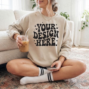 Peut inclure: Sweat-shirt beige avec le texte "YOUR DESIGN HERE" en noir, avec des étoiles et des cœurs. La personne tient un verre de café glacé et un téléphone. La personne porte des chaussettes blanches à rayures noires.