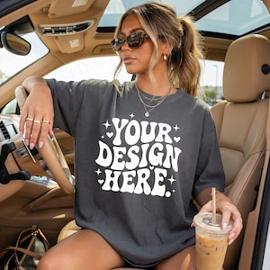 以下が含まれることがあります： 車内で人が着用している、白文字で「YOUR DESIGN HERE」と書かれた濃いグレーのTシャツ。 ゆったりとしたフィット感のシャツです。 サングラスをかけ、飲み物を持っています。