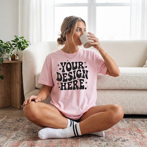 Op de afbeelding: Een lichtroze t-shirt met de tekst "YOUR DESIGN HERE" in het zwart, gedragen door een persoon die met gekruiste benen zit. Het shirt heeft korte mouwen. De persoon houdt een witte mok vast. De achtergrond omvat een witte bank en een patroonkleed.