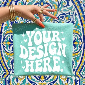 Puede incluir: Una camiseta de color verde azulado claro con el texto "YOUR DESIGN HERE" en blanco, rodeada de estrellas y corazones. La camiseta se muestra sobre un fondo de azulejos coloridos.
