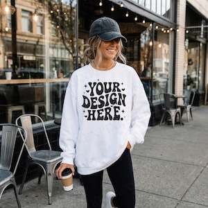 May include: White sweatshirt with the text "YOUR DESIGN HERE" in a black, retro font. The sweatshirt is worn with black leggings and a dark gray baseball cap. A person is holding a coffee cup.