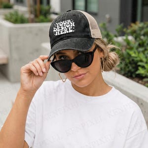 May include: Black and tan trucker hat with the text "YOUR DESIGN HERE" in white. The hat has a curved brim and a mesh back. The person in the image is wearing black sunglasses and a white t-shirt.