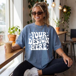 May include: A blue t-shirt with the text "YOUR DESIGN HERE" in white, with star and heart accents. The shirt is worn by a person with sunglasses and jewelry, holding a beverage. The shirt is a relaxed fit.