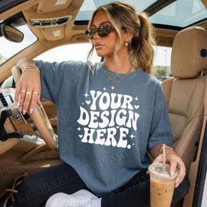 May include: A blue-grey t-shirt with the text "YOUR DESIGN HERE" in white, with star and heart accents. The shirt is worn by a person in a car, holding a drink. The shirt is a casual, oversized style.