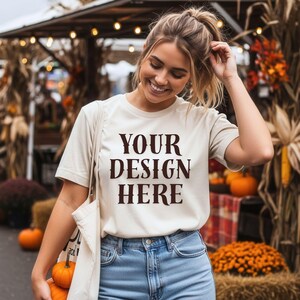 May include: Cream-colored t-shirt with the text "YOUR DESIGN HERE" in a dark brown font. The person is wearing blue jeans and carrying a tote bag. Pumpkins and fall decorations are in the background.