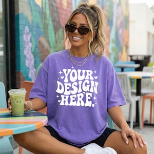 May include: A purple t-shirt with the text "YOUR DESIGN HERE" in white, with a retro font. The person is wearing sunglasses and gold jewelry. The person is sitting at a table with a drink.