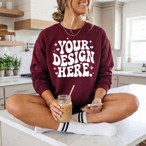 Op de afbeelding: Een bordeauxrode sweatshirt met de tekst "YOUR DESIGN HERE" in het wit, in een retro lettertype. De persoon zit op een wit aanrechtblad en houdt een glazen pot met een rietje en een telefoon vast.
