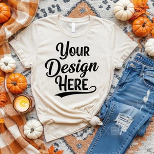May include: Cream-colored t-shirt with the text "Your Design Here" in black script. The shirt is styled with rolled-up sleeves and a knot at the bottom. The shirt is paired with blue jeans and surrounded by pumpkins and fall decor.