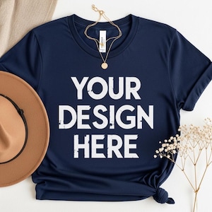 May include: Navy blue t-shirt with the text "YOUR DESIGN HERE" in white. A gold necklace and a brown hat are placed near the shirt. The shirt is a mockup for custom designs.