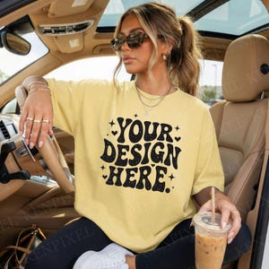 May include: A light yellow t-shirt with the text "YOUR DESIGN HERE" in black, with stars and hearts. The shirt is worn by a person in a car, with sunglasses and jewelry.