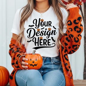 May include: White t-shirt with the text "Your Design Here" in black script, decorated with pumpkin and bat graphics. The person is wearing a burnt orange cardigan with black spots and holding an orange pumpkin mug.